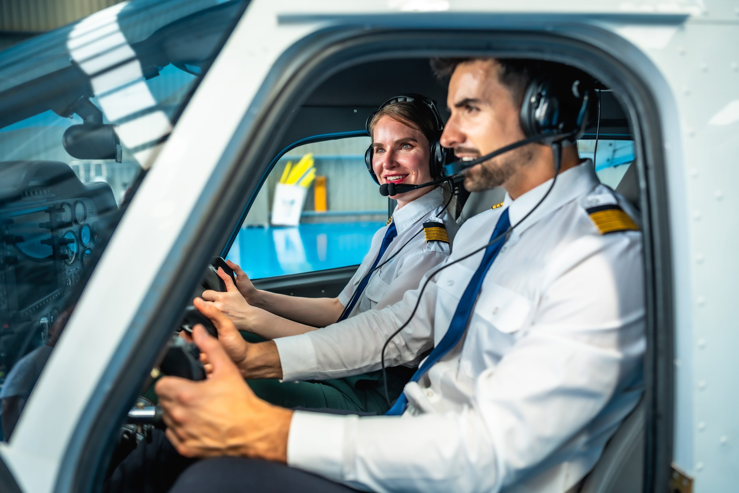 Male and female pilots, wearing headsets and smiling, operating a small airplane inside a hangar, embodying the spirit of aviation